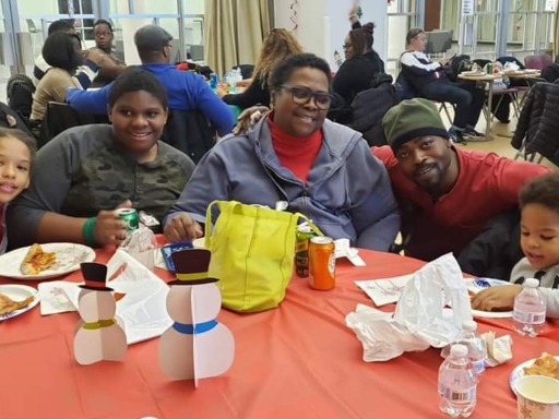 A woman and her family of adults and children sit at a table, sharing a meal at a large event. The woman smiles in a caring manner.