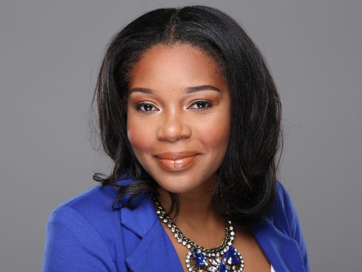 A Black woman poses for a headshot. She is wearing a blue blazer. Grey background.